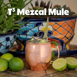 "T3 Mezcal Mule" in a copper mug surrounded by limes and a colorful pot in the background